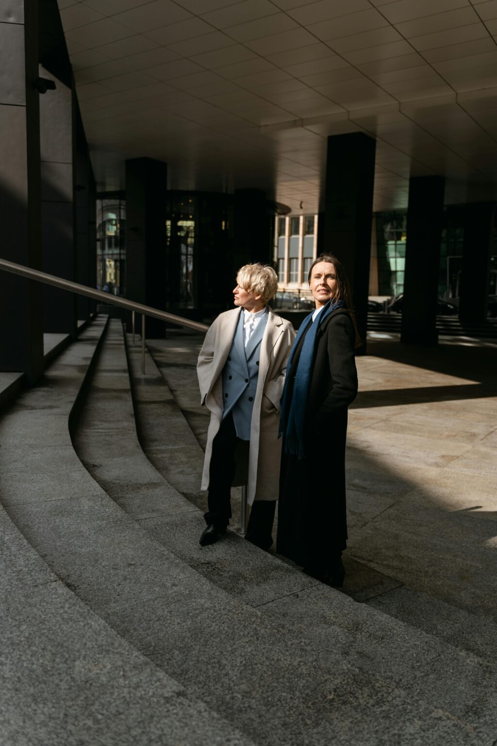 Two businesswomen in formal attire standing on steps outdoors, smiling.
